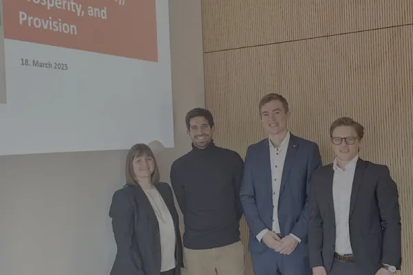 Vier Personen stehen lächelnd vor einer Präsentationsfolie mit der Aufschrift "LAB: Sustainability, Prosperity, and Provision, 18. March 2025". Sie befinden sich in einem modernen Raum mit Holzvertäfelung.