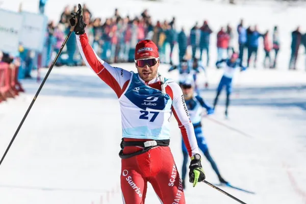 Langläufer überquert bei sonnigem Winterwetter als Sieger die Ziellinie eines Langlaufrennens, trägt einen rot-weißen Rennanzug mit Startnummer 27, hebt den Arm zum Jubel – im Hintergrund jubelnde Zuschauer und schneebedeckte Landschaft.