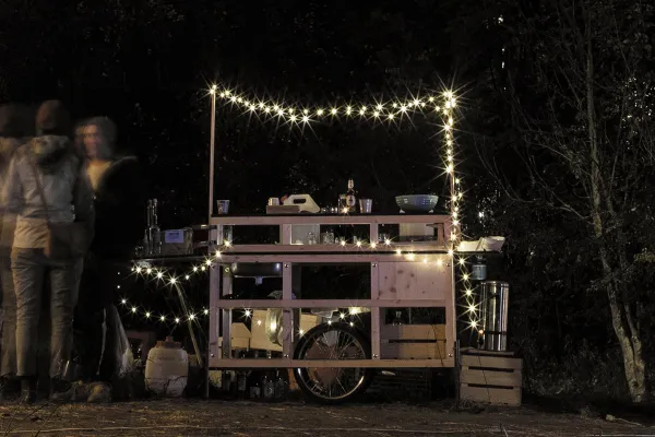 Eine mobile Holzbar, dekoriert mit Lichterketten, steht im Freien bei Nacht. Mehrere Personen stehen links neben der Bar und unterhalten sich. Die Szene vermittelt eine gemütliche und einladende Atmosphäre.
