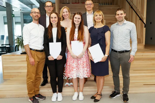 Eine Gruppe von acht Personen steht lächelnd in einem modernen Innenraum. Drei Personen in der vorderen Reihe halten Zertifikate in den Händen. Die Umgebung wirkt hell und einladend mit Holzelementen und Pflanzen.