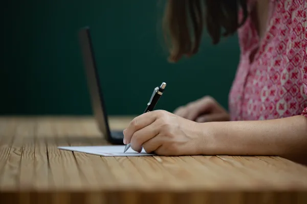 Person schreibt mit Stift auf Papier vor Laptop im Arbeitsraum der Universität Liechtenstein.