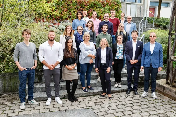 Eine Gruppe von Studierenden und Dozenten des Executive Master of Laws (LL.M.) im Gesellschafts-, Stiftungs- und Trustrecht steht auf einer Treppe im Freien. Sie lächeln in die Kamera, umgeben von grüner Vegetation und einem Gebäude im Hintergrund.