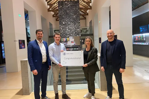 Vier Personen stehen in einem modernen Gebäude und halten ein Schild mit der Aufschrift "Congratulations!" von Bank Frick. Sie feiern die Vergabe eines Stipendiums für den Zertifikatsstudiengang Blockchain und FinTech an der Universität Liechtenstein.