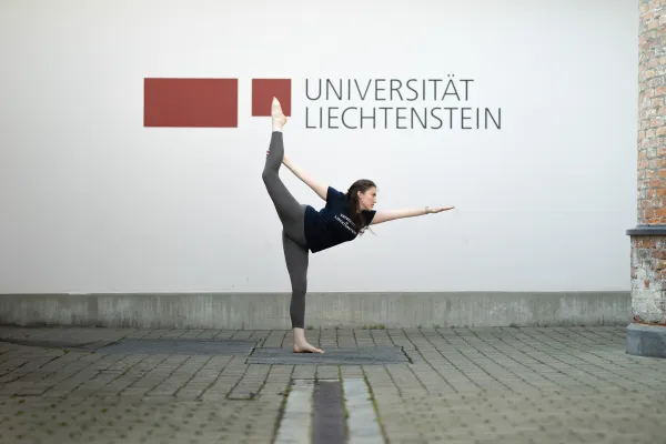 Frau in Sportkleidung übt Yoga-Pose vor Logo der Universität Liechtenstein.