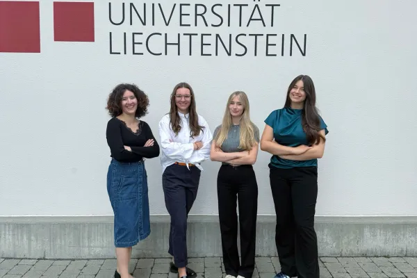 Vier Studentinnen stehen vor der Fassade der Universität Liechtenstein mit Logo und Schriftzug, lächelnd und mit verschränkten Armen, Symbol für Zusammenhalt und Engagement im Studium.