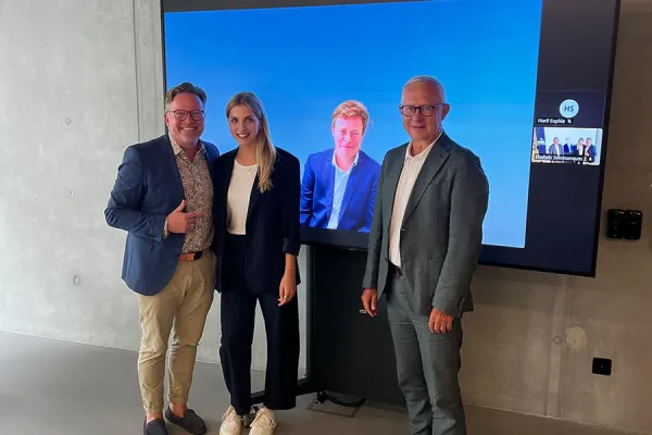 Drei Personen stehen vor einem Bildschirm, der zwei weitere Personen in einem virtuellen Meeting zeigt. Die Szene befindet sich in einem modernen Raum mit Betonwänden.