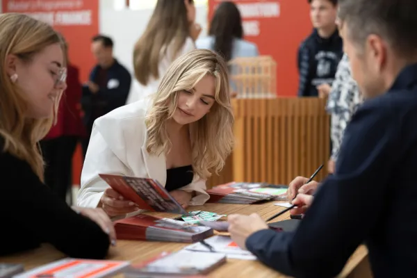 Eine Mitarbeiterin der Universität Liechtenstein berät einen Studieninteressierten an einem Informationsstand und zeigt ihm Informationsmaterialien.