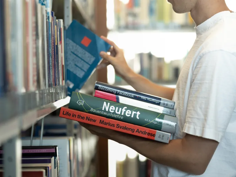 Student wählt Fachliteratur aus dem Bücherregal der Universitätsbibliothek Liechtenstein, mehrere Bücher im Arm.