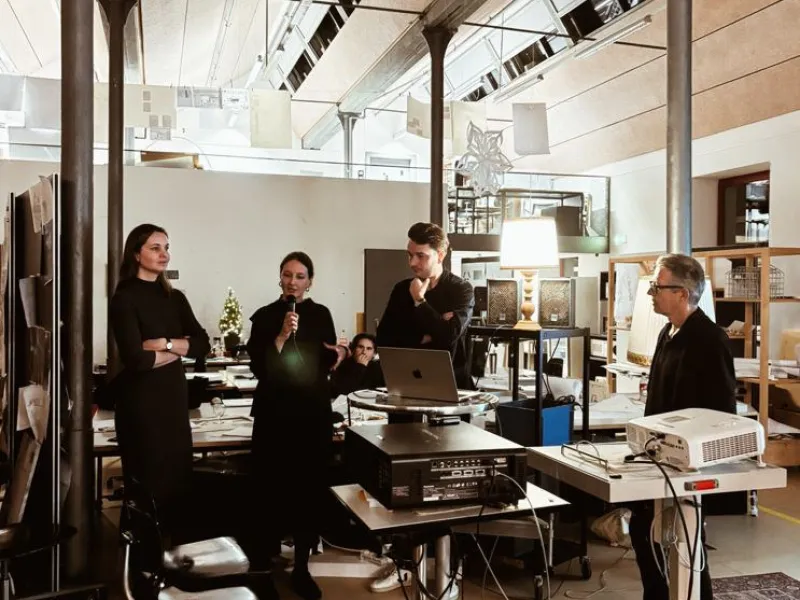 Eine Gruppe von Personen steht in einem modernen Architekturstudio. Eine Person spricht in ein Mikrofon, während andere aufmerksam zuhören. Im Raum sind technische Geräte und Präsentationsmaterialien zu sehen.