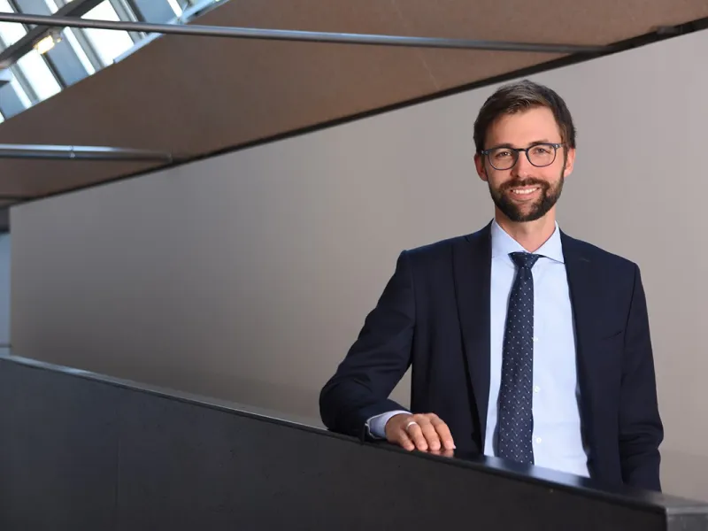 Ein Mann im Anzug steht lächelnd in einem modernen Gebäude mit schrägen Fenstern.