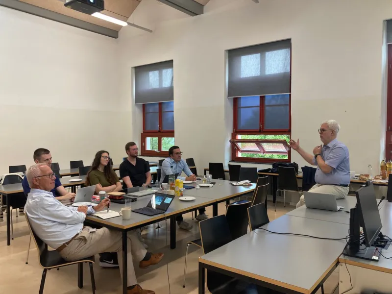 Ein Dozent hält einen Vortrag vor einer kleinen Gruppe von Personen in einem Seminarraum. Die Teilnehmenden sitzen an Tischen mit Laptops und Notizbüchern. Im Hintergrund sind Fenster und eine Leinwand zu sehen.