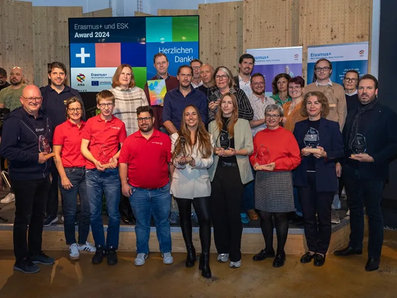 Eine Gruppe von Menschen steht auf einer Bühne und hält Trophäen. Im Hintergrund sind Banner mit Erasmus+ Logos und ein Bildschirm mit der Aufschrift "Erasmus+ und ESK Award 2024" zu sehen. Die Personen lächeln und posieren für ein Gruppenfoto.