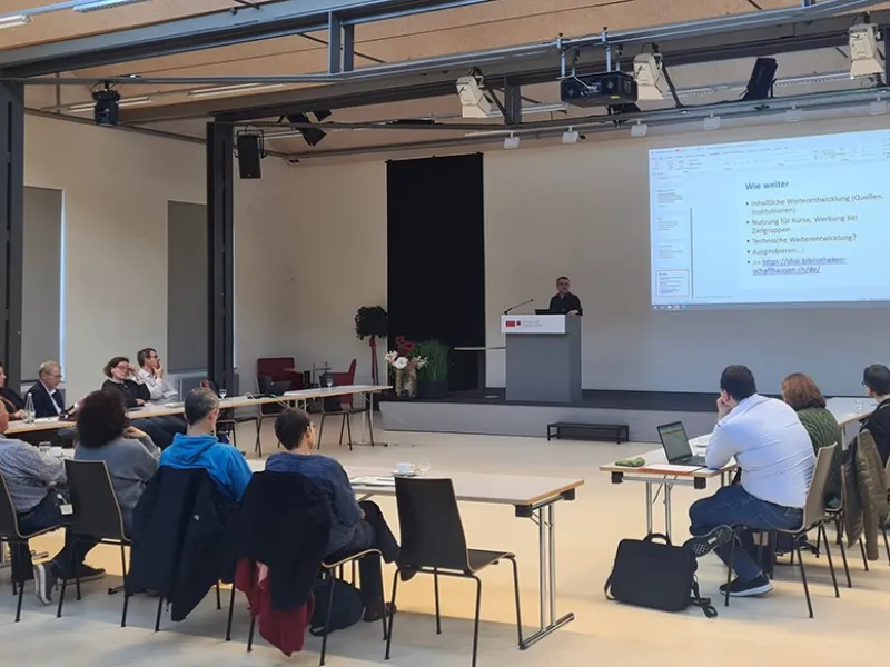 Eine Gruppe von etwa 20 Personen sitzt in einem Auditorium der Universität Liechtenstein, während ein Vortragender an einem Rednerpult spricht. Auf einer großen Leinwand ist eine Präsentation mit dem Titel "Wie weiter" zu sehen.